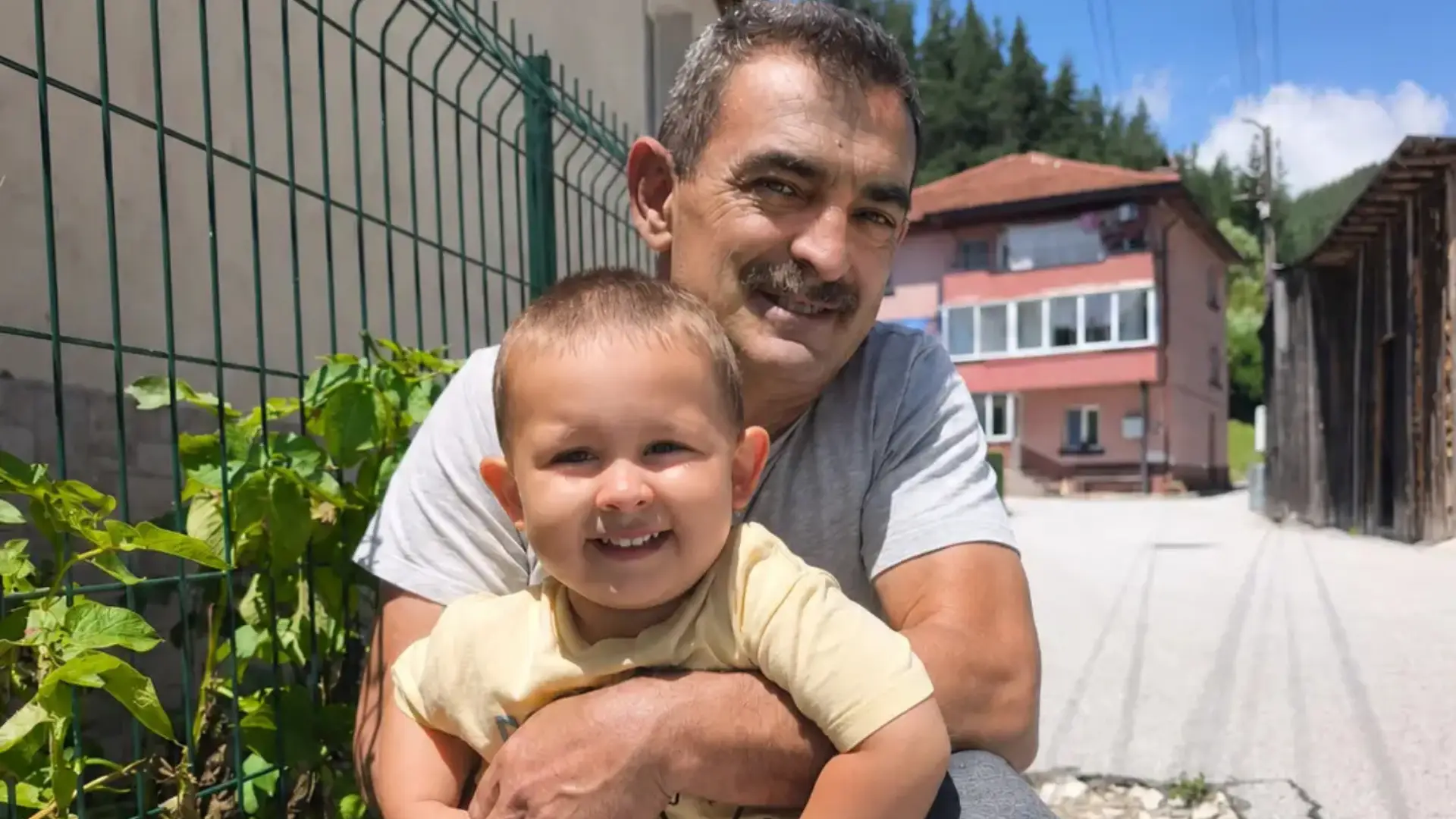 Снимка на щастлив мъж на средна възраст, клечащ на улицата и държащ в обятията си усмихнатото си малко внуче, правена в по-добри дни, преди парализата.